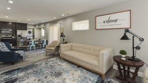 Een moderne woonkamer met een beige bank, een blauwe fauteuil, een salontafel en een vloerkleed. Op de achtergrond is een eethoek met een tafel en stoelen.