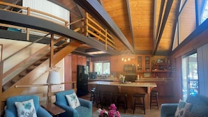 A spacious living area with a wooden staircase, a kitchen with a bar, and a dining area with blue chairs. - Lake Almanor Peninsula Cabin In The Pines (Westwood)
