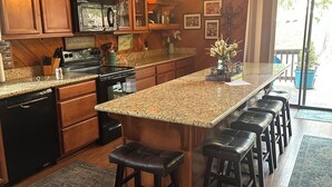 A kitchen with wooden cabinets, a granite countertop, a central island with bar stools, and a large window. - Lake Almanor Peninsula Cabin In The Pines (Westwood)