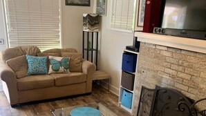 Een woonkamer met een beige bank, twee blauwe kussens in de vorm van vlinders, een glazen salontafel met blauwe onderzetters, een open haard met bakstenen omlijsting, en een flatscreen televisie op een wit plank.