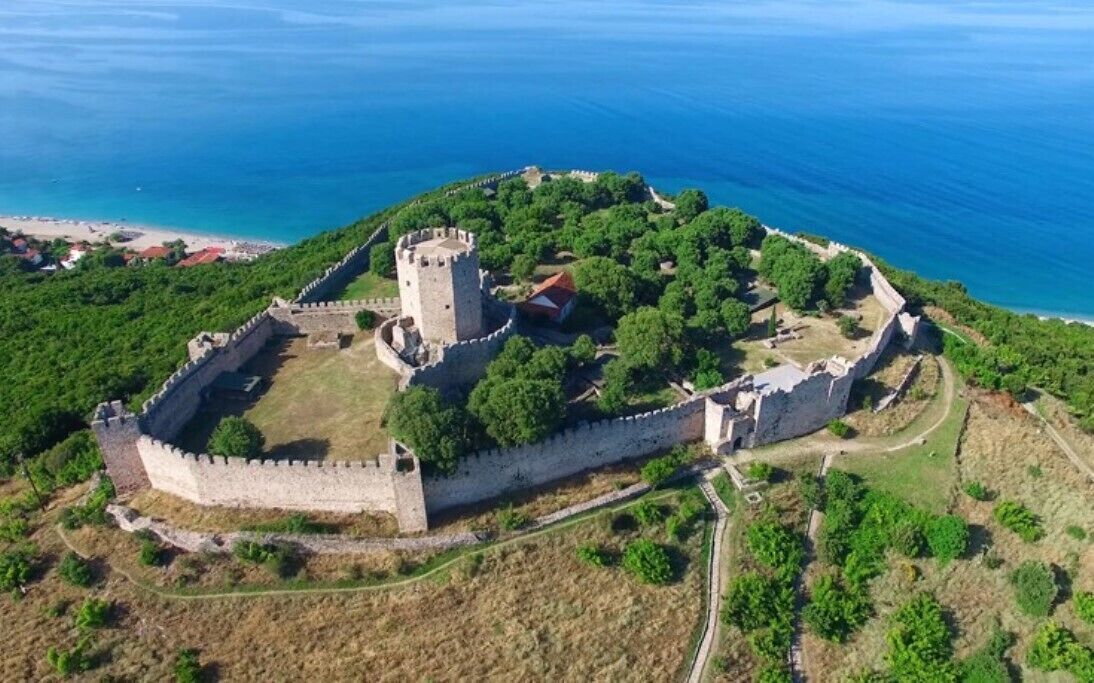 An aerial view of a historic castle surrounded by a fortified wall, with a tower and a red-roofed building, overlooking a coastal area.