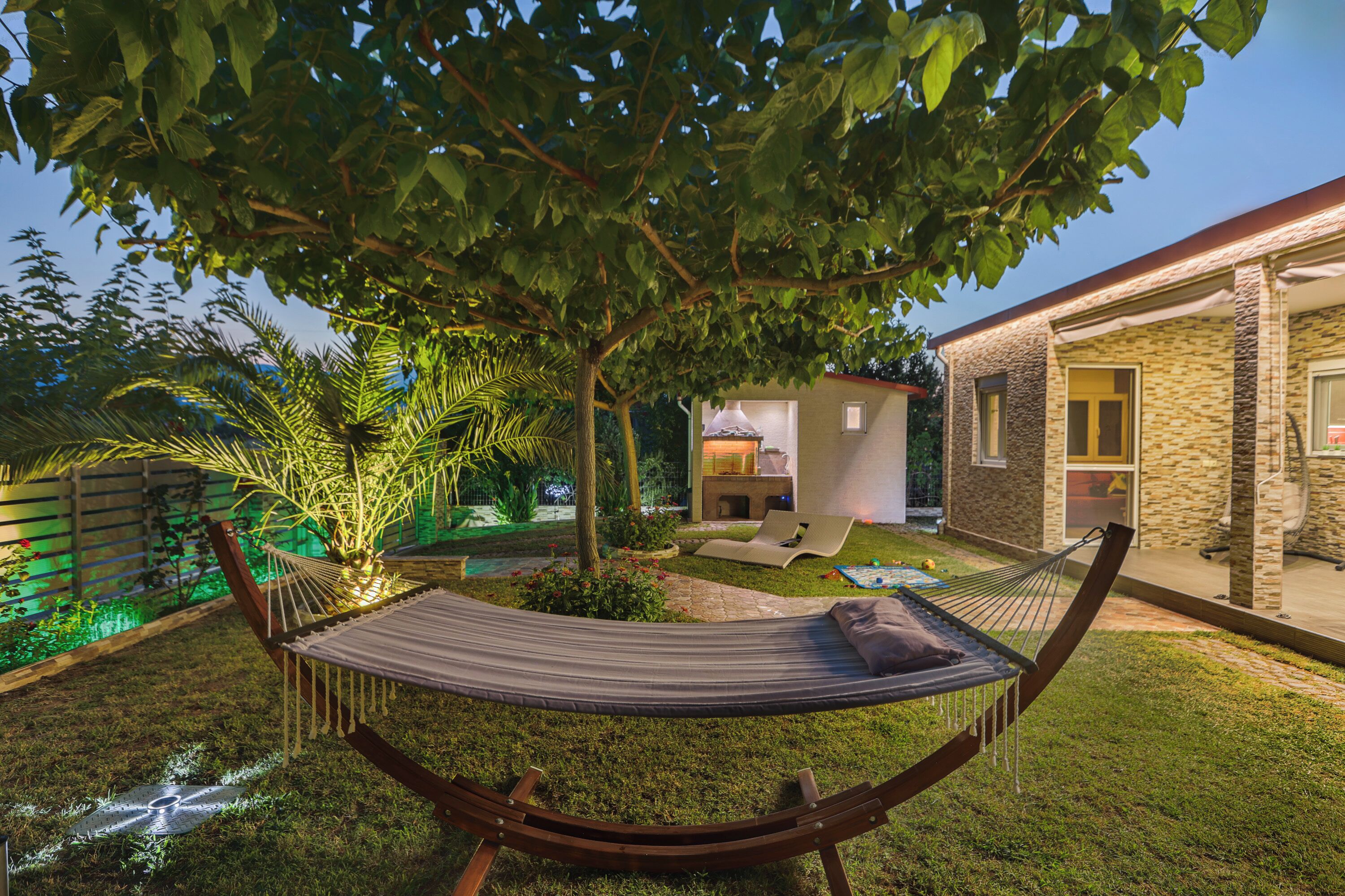 Un hamac suspendu sous un arbre, dans un jardin bien entretenu, avec un bâtiment en pierre en arrière-plan.