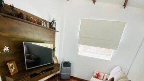 Ein Wohnzimmer mit beiger Couch, einem Flachbildfernseher an einer Holzwand, einem kleinen Heizgerät und einem Fenster mit Jalousien.