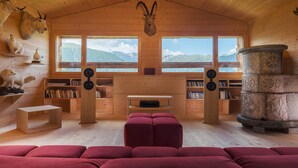 A cozy cabin interior with wooden walls, a large window overlooking mountains, a stone fireplace, and a collection of stuffed animals on shelves. - Exclusive architectural chalet at 1,600 metres, 4-6 pers. in impressive location (Suen)