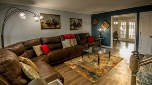 Een woonkamer met een bruine leren bank, een glazen salontafel en een kleurrijk vloerkleed. Er hangen schilderijen aan de muur en een trap die leidt naar een andere kamer.