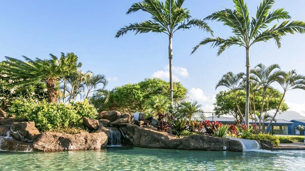 Un espace piscine d’un complexe hôtelier agrémenté d’une cascade, de palmiers et d’un bâtiment en arrière-plan.