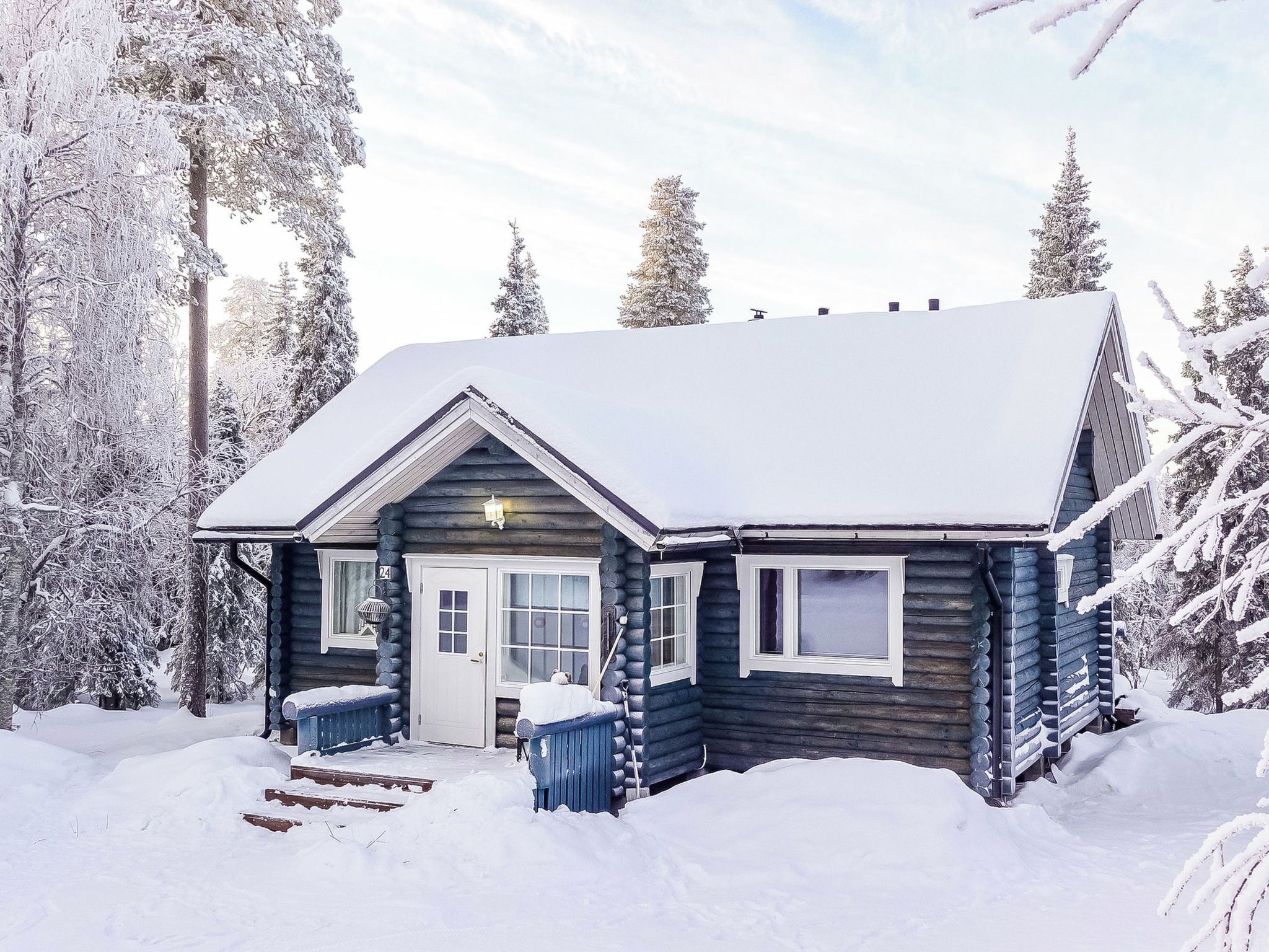 A log cabin with a snow-covered roof, a front porch, and a visible number '24' on the door.