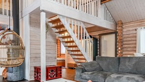 Un vaste espace de vie comprenant un canapé gris, une table basse en bois et un fauteuil en osier. Un escalier mène à un niveau supérieur.