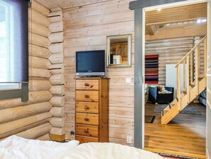 A log cabin interior with a bed, wooden dresser, television, and a staircase leading to another room.