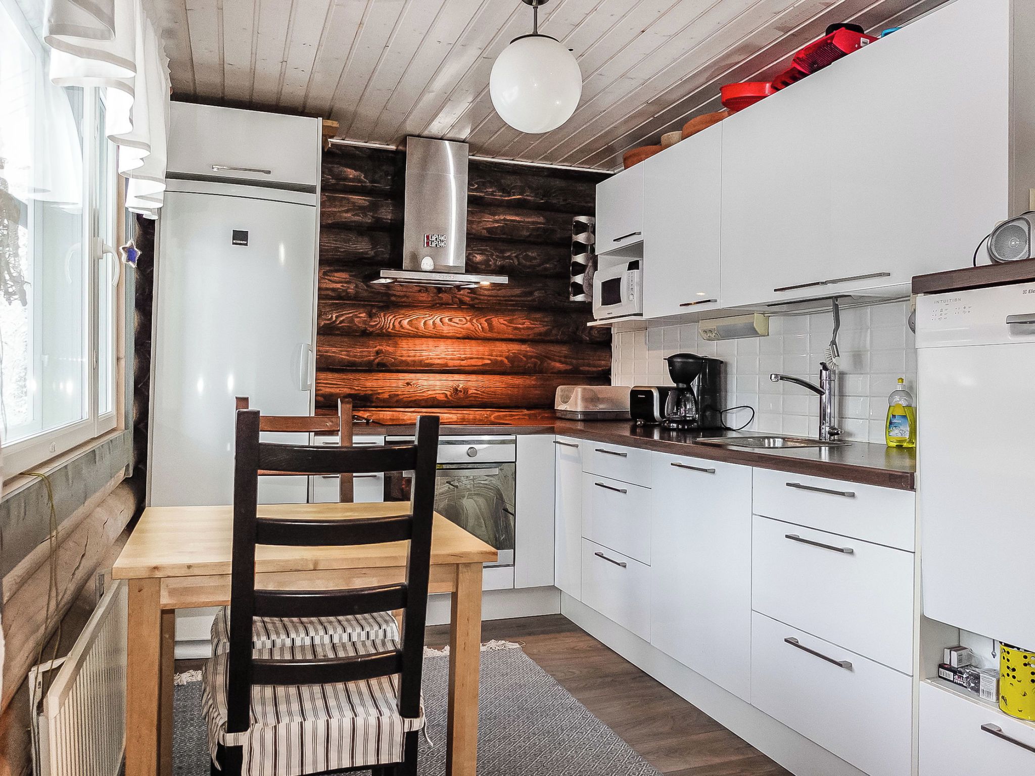 Cocina compacta con armarios blancos, una pared decorativa de madera, una mesa de comedor con una silla y una lámpara colgante.