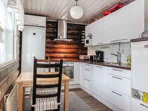 A compact kitchen with white cabinets, a wooden accent wall, a dining table with a chair, and a hanging light fixture. - Isopyhänmaa by Interhome (Salla)
