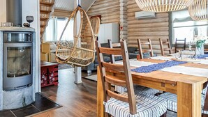 L’intérieur chaleureux d’un chalet avec une grande table à manger en bois placée au centre, des chaises en osier suspendues, une cheminée en pierre et de grandes fenêtres laissant entrer la lumière naturelle.