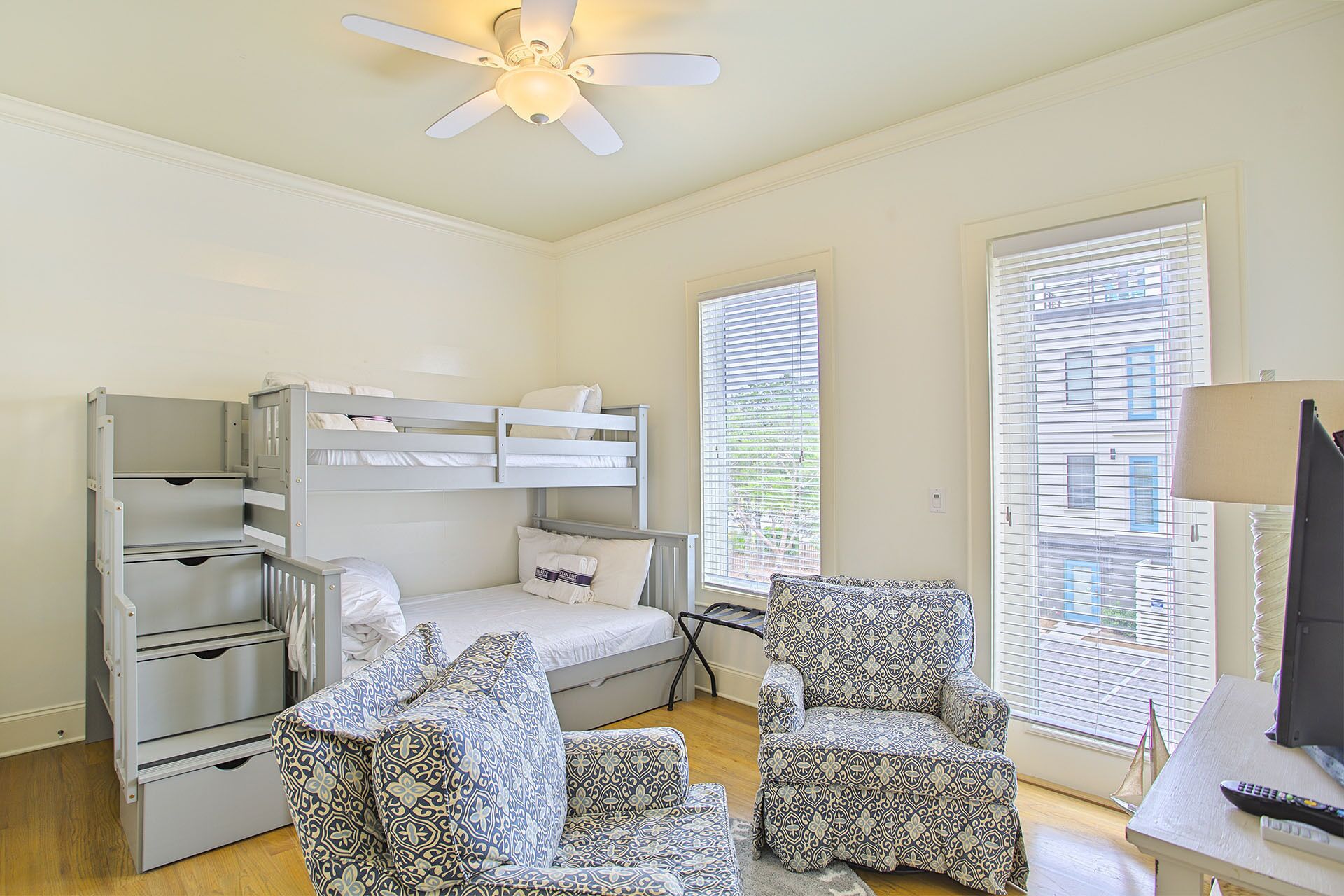 A bedroom with bunk beds, a desk, two patterned chairs, a ceiling fan, and large windows with blinds.