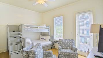 A bedroom with bunk beds, a desk, two patterned chairs, a ceiling fan, and large windows with blinds.