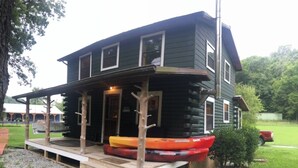 A log cabin with a porch, a small deck, and kayaks stored outside.