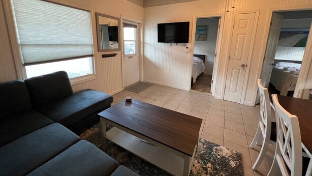 Un espacio habitable compacto con sofá, una mesa de centro, un televisor montado en la pared y una puerta que da a un dormitorio.