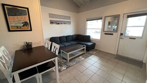 Una sala acogedora con una mesa de comedor marrón oscuro, sillas blancas, un sofá gris y una mesita de centro. Hay un cuadro en la pared y un letrero sobre el sofá.
