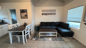 Un salón con un sofá oscuro, una mesa de centro y una mesa de comedor con sillas blancas. Hay un cartel en la pared que dice "POINT PLEASANT BEACH".