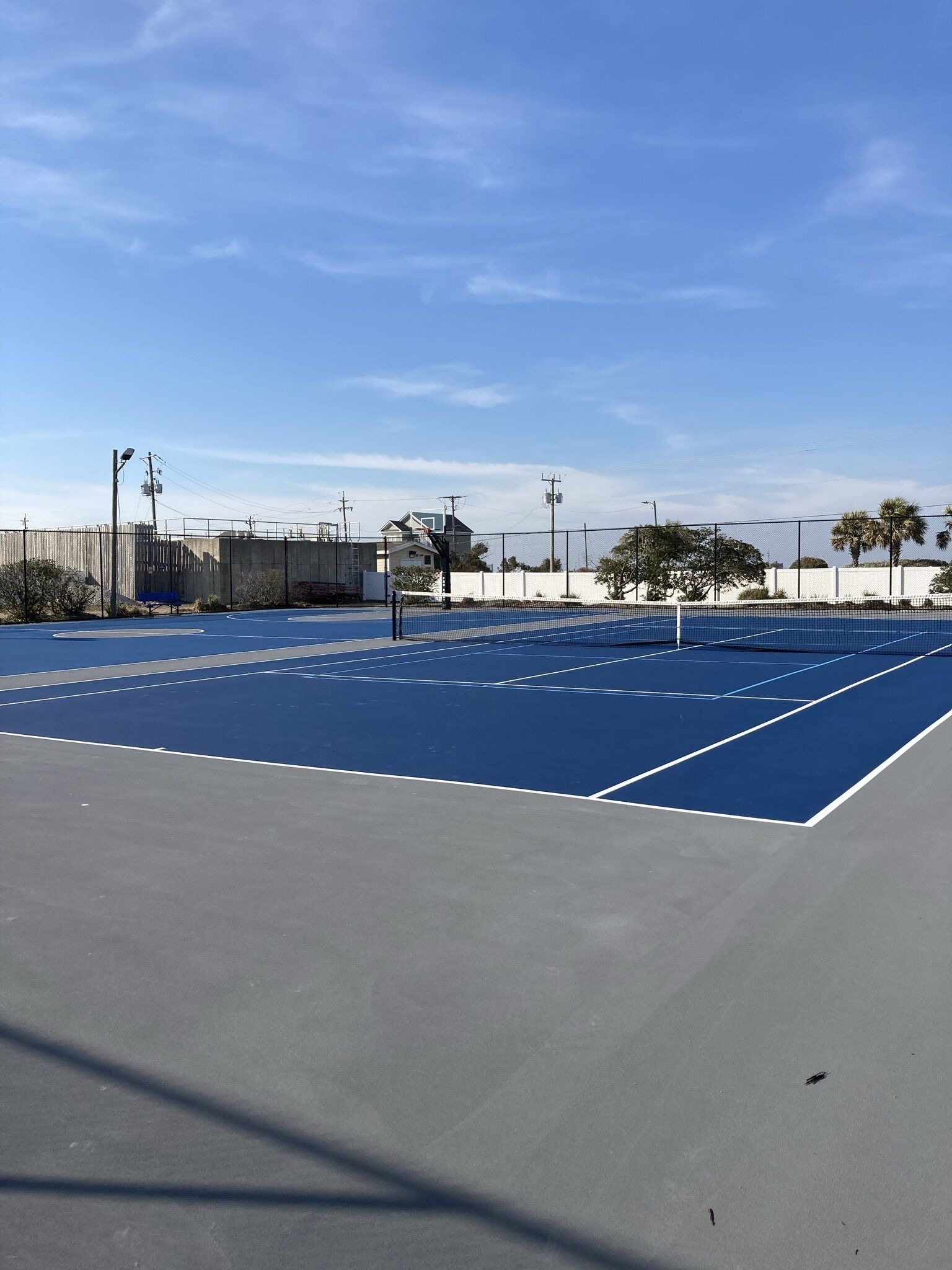 An outdoor tennis court with a blue surface, surrounded by a fence, and utility poles in the background.