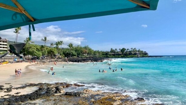 A beach with clear turquoise water, people swimming and walking on the shore, and a backdrop of palm trees and a building. - Kona Ocean View:  Beautifully Furnished, Amazing View, White Sands Beach (Kailua-Kona)