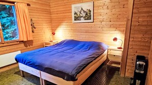 Una habitación de cabaña de madera con cama, dos mesitas de noche con lámparas, una ventana con cortinas, un cuadro en la pared y un calentador en el suelo.