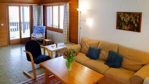 Una sala acogedora con piso de madera, un sofá color beige, una mesa de centro de madera y una silla. Hay una ventana con cortinas, una lámpara y un cuadro en la pared.
