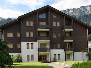 Un edificio de varios pisos con balcones, una entrada central y montañas al fondo.