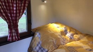 Una habitación con una cama cubierta por un edredón amarillo estampado en cuadros, una ventana con cortinas de cuadros rojos y blancos, y una lámpara fijada a la pared.