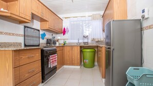 Eine Küche mit Holzausstattung, einem Edelstahlkühlschrank, einer grünen Mülltonne und einem Fenster mit durchsichtigen Vorhängen.