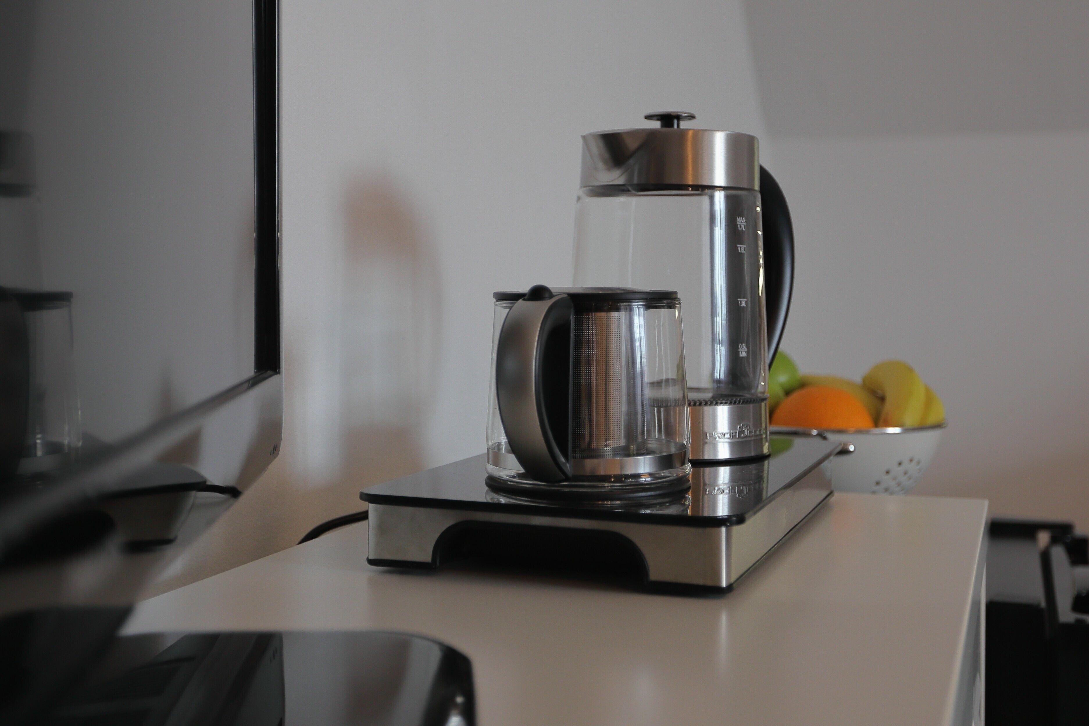 Una cafetera sobre una encimera con un frutero al fondo.
