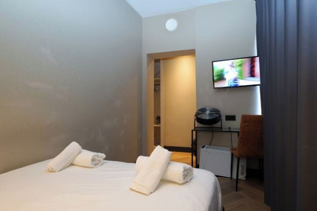 A hotel room with a bed, two rolled towels, a television on the wall, a desk with a chair, and a small heater.