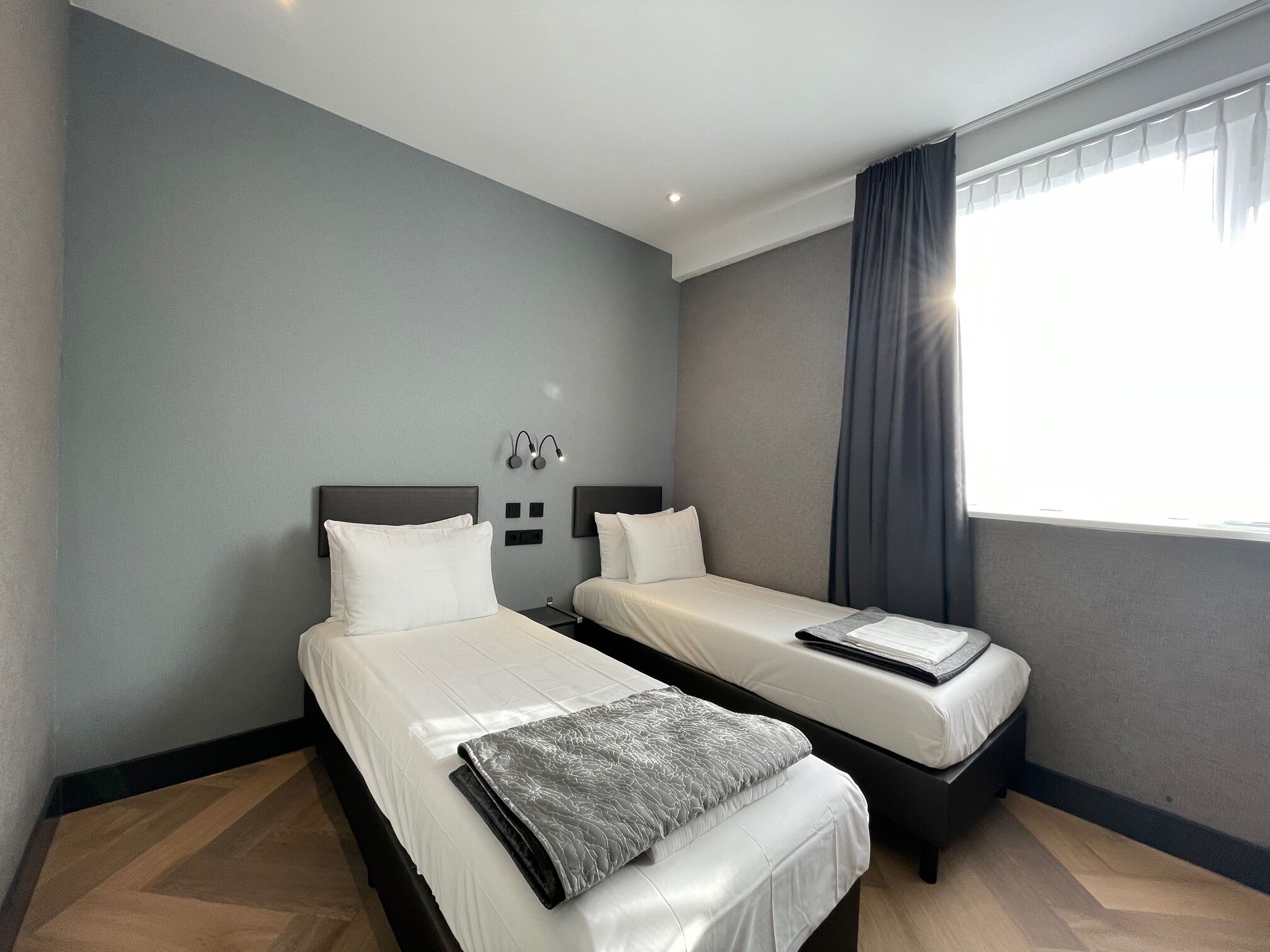 A hotel room with two single beds, a small table, a wall-mounted light fixture, and a window with curtains.