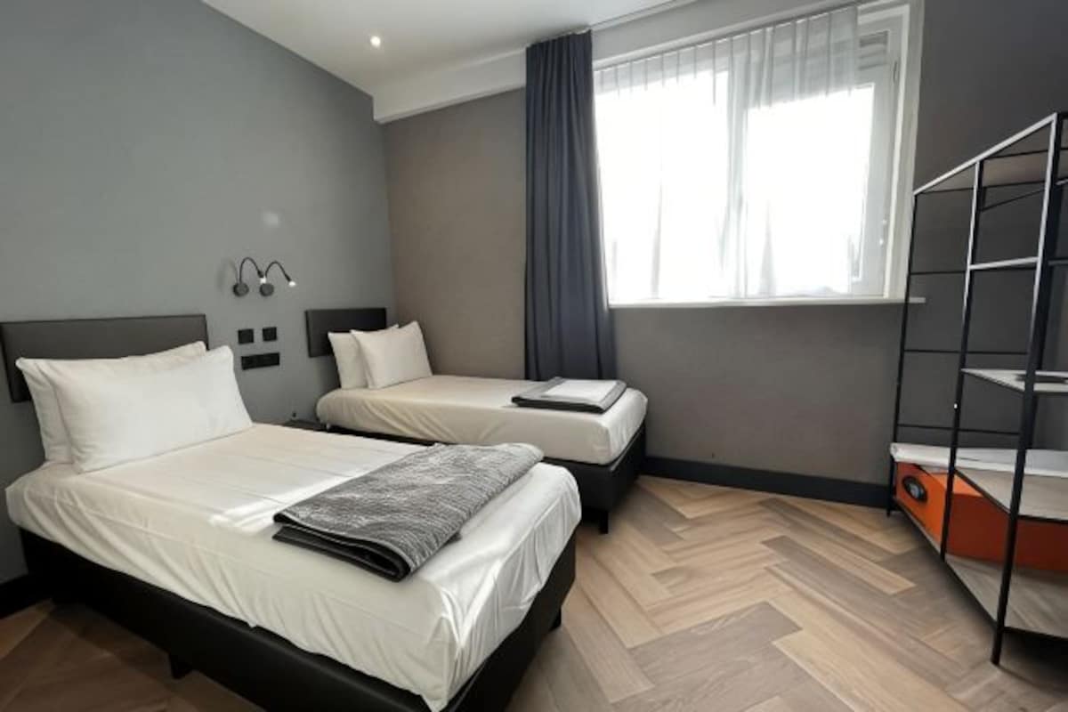 A hotel room with two beds, a window with curtains, a wooden floor, and a metal shelf unit.