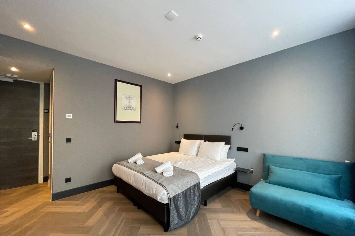 A modern bedroom with a bed, a teal sofa, a framed artwork, and a wooden floor.