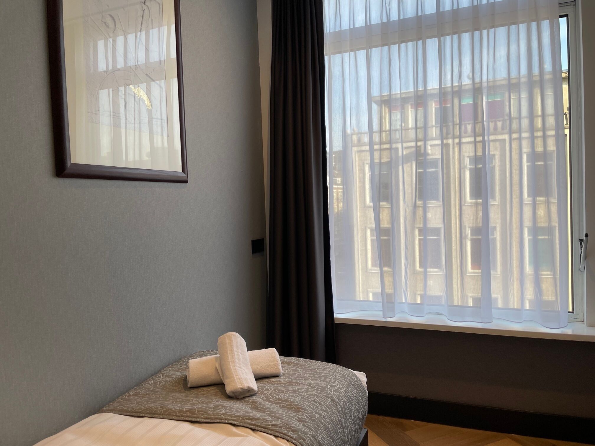 A hotel room with a bed, a window with sheer curtains, and a framed picture on the wall.