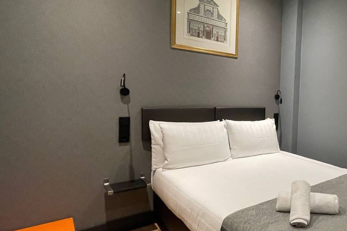 A hotel room with a bed, a nightstand, a framed picture on the wall, and a small orange box on the floor.