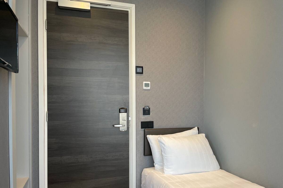A hotel room with a wooden door, a bed with white bedding, a nightstand, and a wall-mounted control panel.