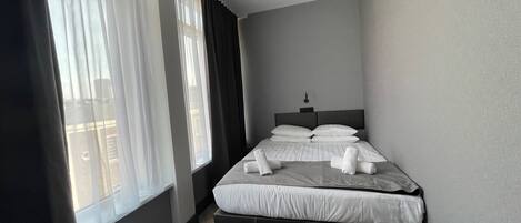 A neatly made bed with white linens and towels, a bedside table with a lamp, and a window with sheer curtains.