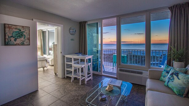Ein Balkon mit Blick auf den Sonnenuntergang über dem Meer, ein weißes Sofa, ein kleiner Tisch und ein Stuhl.