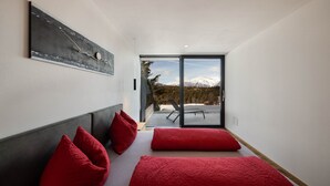 Un dormitorio moderno con una cama grande, almohadas rojas y un ventanal que da a un balcón con vistas a la montaña.