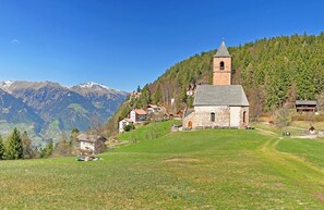 A small stone church with a bell tower, surrounded by a green field and houses, with mountains in the background.