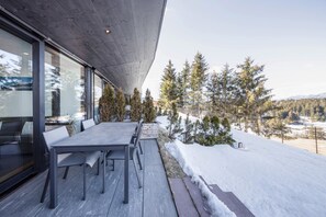 Ein moderner Essbereich im Freien mit Betonüberdachung, einem langen Tisch und Stühlen, mit Blick auf eine verschneite Landschaft mit Bäumen und Bergen.