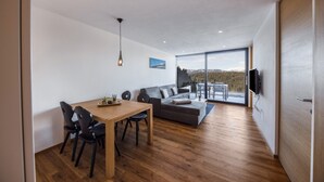 Ein modernes Wohnzimmer mit Holzboden, einem Esstisch mit Stühlen, einem grauen Sofa, einem an der Wand montierten Fernseher und einem großen Fenster mit Blick ins Freie.