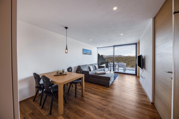 Ein modernes Wohnzimmer mit Holzboden, einem Esstisch mit Stühlen, einem grauen Sofa, einem an der Wand montierten Fernseher und einem großen Fenster mit Blick ins Freie.