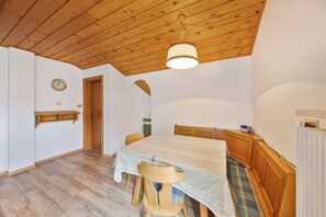 Ein Zimmer mit Holzbalkendecke, einem Bett mit Kopfteil, einem Holzstuhl, einem Tisch mit Tischdecke und einem kleinen Regal.