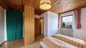 A bedroom with a wooden ceiling, a window with a view, a bed with patterned bedding, and a door leading to another room.