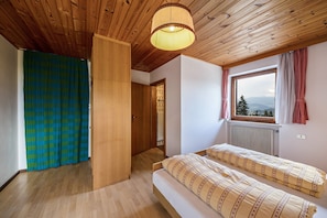 Een slaapkamer met een houten plafond, een raam met uitzicht, een bed met beddengoed met een patroon en een deur naar een andere kamer.