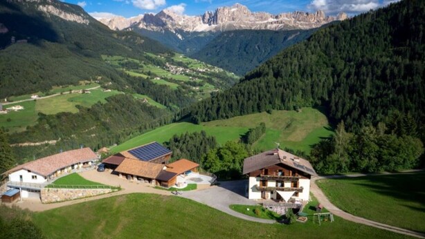 A rural mountain village with traditional houses, a solar panel installation, and a valley with mountains in the background.