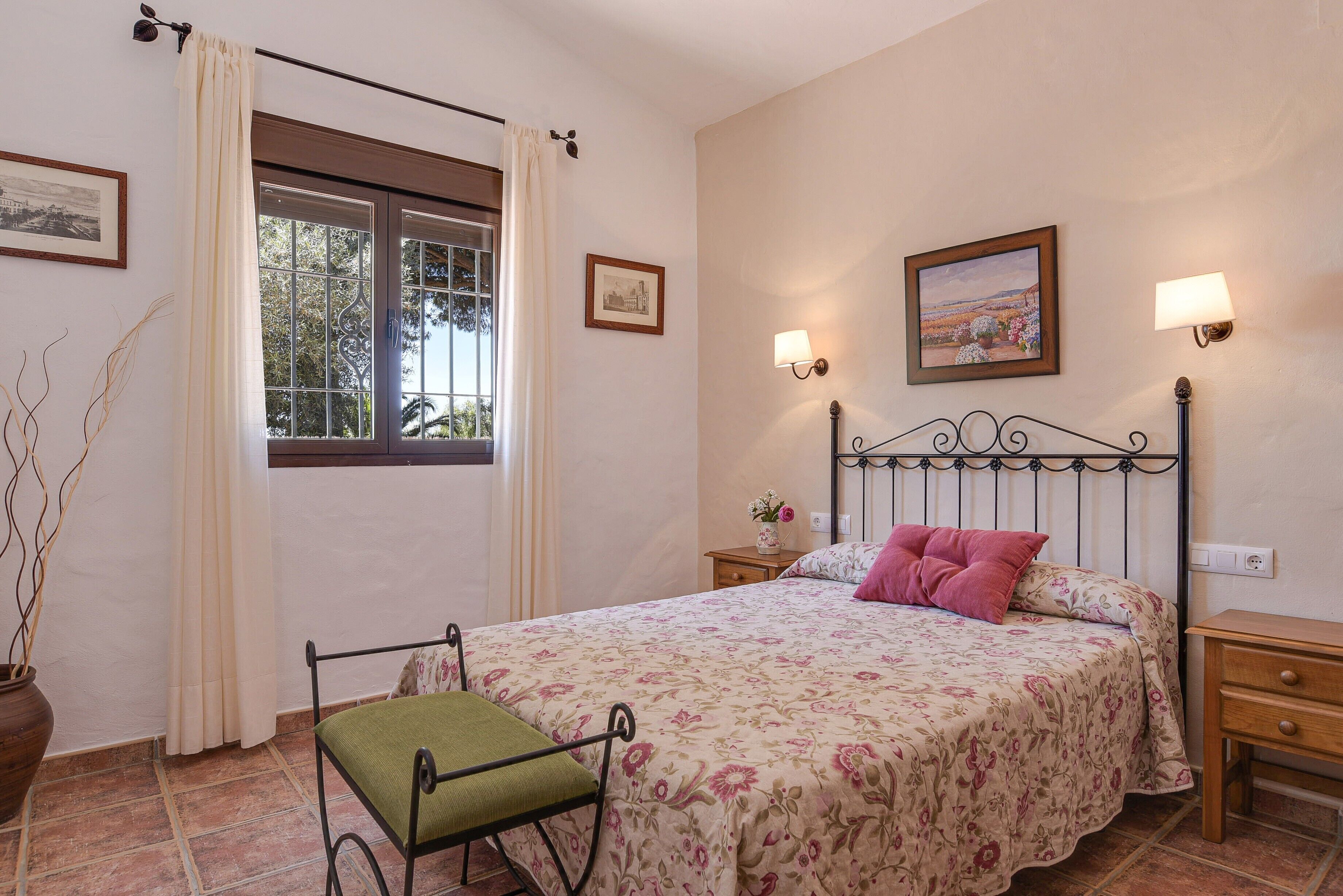 Un dormitorio con cama, mesita de noche, planta en maceta y lámparas de pared. Tiene cortinas en la ventana.