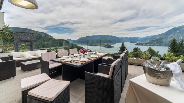 Ein Essbereich im Freien mit Korbmöbeln, einem Tisch für vier Personen und Blick auf einen See und Berge.